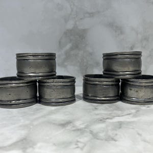 May include: Six antique silver-toned napkin rings. Each ring is cylindrical with a slightly textured surface and two raised bands. The rings are arranged in two stacks of three, suggesting a set for a dining table.
