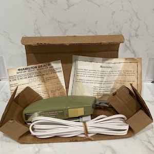 May include: An open cardboard box containing a vintage Hamilton Beach electric knife. The olive green knife has a white cord and is accompanied by product information and a guarantee card. The box is partially open, revealing the contents.