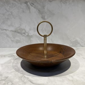 May include: A round wooden serving bowl with a smooth, dark brown grain finish. A vertical brass-toned metal post rises from the center, topped with a circular brass-toned ring handle, creating a tiered-style serving piece for food or items.