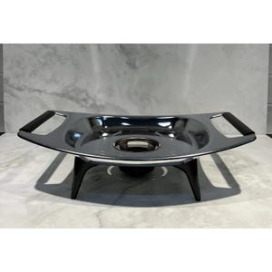 May include: A silver-colored metal fondue stand with a polished finish, featuring two black handles on the sides and three black legs. The center of the stand has a circular opening designed to hold a heat source for warming food.