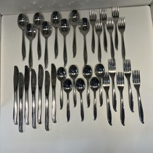 May include: A large collection of polished stainless steel flatware, including various sizes of spoons, forks, and knives. Each piece features a smooth, reflective finish and a simple, curved handle design, arranged in rows on a plain surface.