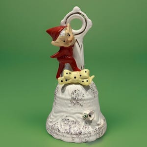 May include: A vintage ceramic bell featuring a whimsical elf figure in a red outfit and hat. The bell is white with a textured surface and a yellow bow with black spots. A decorative piece with a festive theme.
