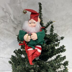 May include: A Christmas elf ornament with a red hat and boots, green jacket, and white beard. The elf is holding a gold bell and is perched on a small Christmas tree. The elf's legs are striped red and white.