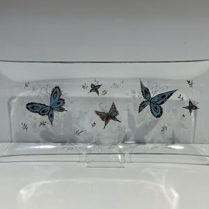 May include: A rectangular clear glass serving tray with a curved edge. The tray is decorated with blue and brown butterfly designs and small floral accents. The tray is supported by a clear plastic stand.