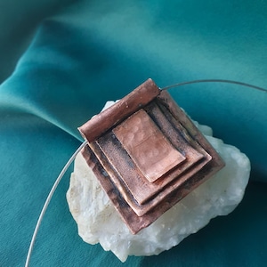 Puede incluir: Un collar con colgante de cobre con un diseño cuadrado en capas. El colgante tiene un acabado rústico y oxidado y está suspendido de un fino alambre plateado. El collar se exhibe sobre una piedra blanca sobre un fondo turquesa.