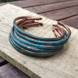 Könnte beinhalten: Ein Stapel Kupferarmbänder mit türkisfarbener Patina. Die offenen Armreifen sind auf einer verwitterten Holzoberfläche gestapelt. Die Armbänder haben ein rustikales, gealtertes Aussehen mit sichtbaren Farb- und Texturvariationen.
