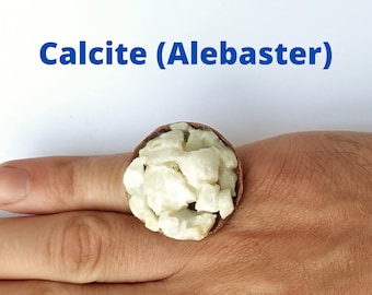 May include: A ring featuring a copper-colored band holding a cluster of white calcite (alabaster) stones. The text "Calcite (Alebaster)" is displayed in blue above the ring. The ring is shown on a person's hand.