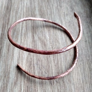 May include: A handcrafted copper bracelet with a hammered texture. The spiral design features a warm, reddish-brown hue, and the ends are slightly curved. This unique piece of jewelry is designed to be worn on the wrist.
