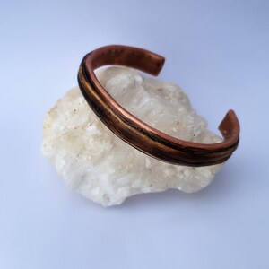 Puede incluir: Una pulsera de puño de cobre con incrustaciones marrones oscuras y doradas. La pulsera está abierta y descansa sobre una piedra blanca texturizada. El metal tiene un tono cálido marrón rojizo.