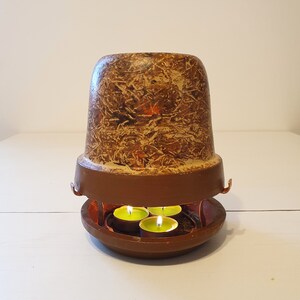 May include: A terracotta candle heater with a brown base and a textured, decorative top. The base holds three lit tealight candles, creating a warm glow. The design is rustic and suitable for home decor. The heater is a charming addition.