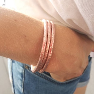 May include: Three hammered copper bangles on an arm. The bracelets have a textured surface and a warm, rose-gold color. The person is wearing a white shirt and blue denim shorts.