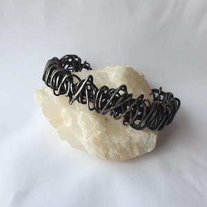 Può includere: Un braccialetto in metallo grigio scuro con un design a filo intrecciato e aggrovigliato. Il braccialetto è appoggiato su una pietra chiara e strutturata. Il design del braccialetto presenta una serie di anelli e vortici interconnessi, creando un aspetto artistico unico.