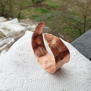 May include: A handcrafted copper bracelet with a curved, open design. The bracelet has a polished finish and is displayed on a white, textured surface. The warm, reddish-brown colour of the metal contrasts with the background.