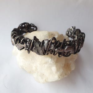 Può includere: Un bracciale a polsino argentato fatto a mano con un design astratto. Il bracciale presenta un'estetica strutturata e organica con filo intrecciato e forme irregolari. Il metallo ha una finitura scura e ossidata, che gli conferisce un aspetto vintage o industriale. Il bracciale è esposto su una superficie bianca.