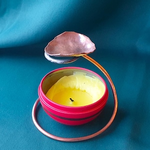 May include: A handmade candle holder with a copper-colored metal frame. The holder features a small, round, red tin with a yellow candle inside. A copper-colored dish sits above the candle, and the base is a copper-colored ring.