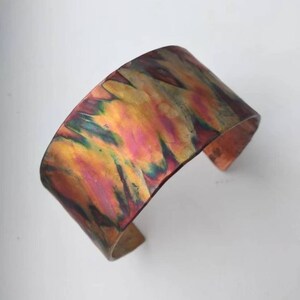 Puede incluir: Pulsera ancha de cobre con superficie texturizada. La pulsera presenta un vibrante patrón iridiscente de tonos amarillos, rosas, verdes y bronce. El diseño abierto facilita su uso y añade un toque artístico.