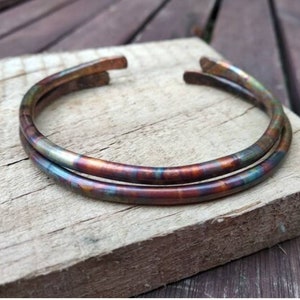 Puede incluir: Dos pulseras abiertas de color cobre con un acabado iridiscente. Las pulseras, de forma circular, muestran una mezcla de tonos rojos, verdes y azules, sobre una superficie de madera envejecida.