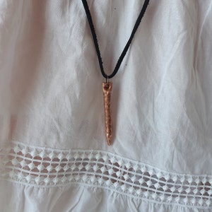 Può includere: Una collana con pendente color rame con un design allungato e strutturato. Il pendente è appeso a un cordino nero. La collana è esposta su uno sfondo bianco.