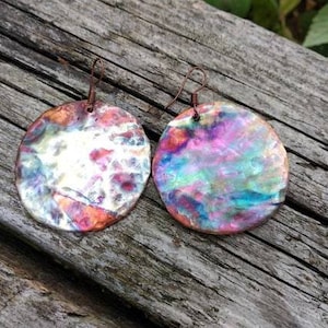 Może przedstawiać: Para ręcznie robionych, okrągłych kolczyków z kolorowym, opalizującym wykończeniem. Kolczyki posiadają miedziany druciany haczyk i unikalny, abstrakcyjny wzór z odcieniami różu, błękitu, fioletu i pomarańczy. Na tle rustykalnego drewna.