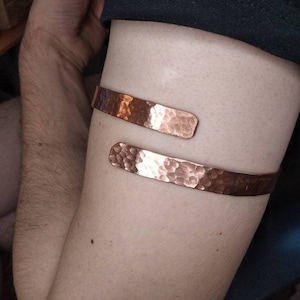 Puede incluir: Un brazalete de cobre martillado con una superficie texturizada. El brazalete es de un color marrón rojizo cálido y se envuelve alrededor del brazo. El brazalete está abierto en la parte delantera y tiene un diseño simple y minimalista.