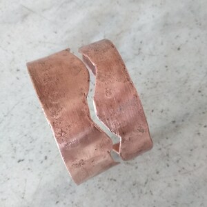 Può includere: Un bracciale a polsino color rame con finitura anticata. Il bracciale è aperto, con un'apertura al centro. Il metallo ha un aspetto strutturato e i bordi sono leggermente irregolari.