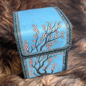 Op de afbeelding: Een blauwe doos met scharnieren en een kersenbloesemontwerp. De doos heeft een lichtblauwe achtergrond met een gedetailleerde illustratie van een kersenbloesemboom in volle bloei. De randen zijn donkerbruin met decoratieve stiksels.