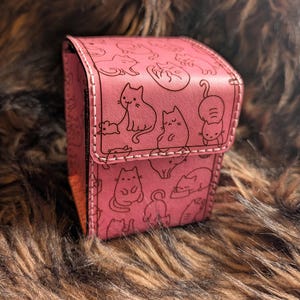May include: A pink rectangular storage box featuring a repeating black line art pattern of various cats in different poses. The box has a hinged lid with visible white stitching along the edges and a smooth, matte finish on the exterior surface.
