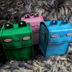 Puede incluir: Tres cajas hechas a mano con forma de rana en verde, rosa y azul. Cada caja presenta detalles cosidos, incluyendo ojos, boca y un contorno cosido. Las cajas están sobre una superficie texturizada y peluda.
