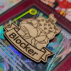 May include: A wooden "Blocker" emblem rests on a trading card featuring Eustass "Captain" Kid. Other cards with colourful illustrations are visible, including one with Shirahoshi. The cards are in a red binder.