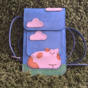 May include: A blue leather crossbody bag with a pink cat and clouds design. The bag has a flap closure and a long adjustable strap.