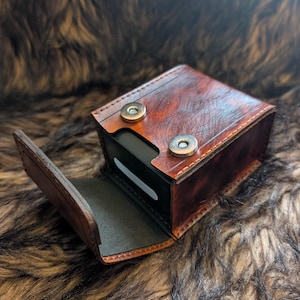 May include: A rectangular brown leather case with a marbled finish and visible stitching along the edges. The case features a hinged lid secured by two circular silver-toned metal magnetic snaps, revealing a black interior compartment.