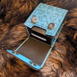 May include: A light blue deck box featuring a black line art pattern of various cats. The box has a fold-over flap secured by two silver-toned metal snap buttons, with a black interior lining and a rectangular white slot on the front panel.