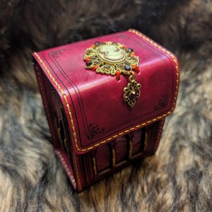 May include: A deep magenta leather box with gold stitching and black line detailing. The lid features an ornate gold-tone metal brooch with a central cameo and dark crystals, with a dangling gold-tone metal charm attached to the bottom.