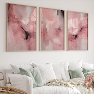 May include: A set of three abstract art prints. Each print features a pink and white abstract design with black accents. The prints are framed in a light wood frame.