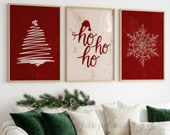 May include: Three framed wall art prints featuring holiday themes. The left print shows a white scribbled Christmas tree on a red background. The center print displays red text reading ho ho ho with a Santa hat on a cream background. The right print features a white snowflake on a red background.