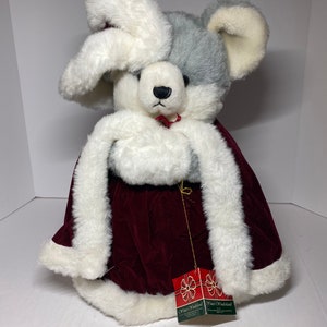 May include: A plush gray and white mouse wearing a red velvet dress with a white fur trim and a white fur hat. The mouse is holding a red bow in its paws. The mouse has a tag that says "Winter Wonderland".