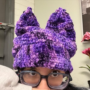 May include: Purple and black fuzzy knit hat with ear flaps. The hat is made of a soft, fluffy material and has a unique, textured design.