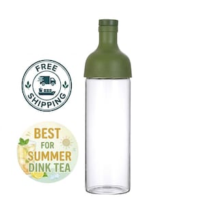 May include: A clear glass bottle with a cylindrical body and a matte olive green cap and neck section. The bottle features a smooth, transparent glass base and a solid-colored top component that tapers to a narrow opening with a matching green lid.