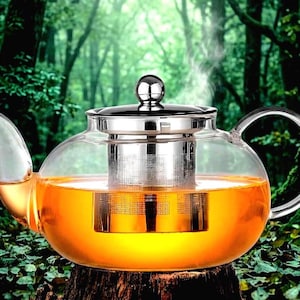 May include: A clear glass teapot with a stainless steel infuser and lid, filled with amber-colored tea. The teapot is steaming, suggesting hot tea. The handle and spout are also clear glass. The teapot is sitting on a wooden surface.