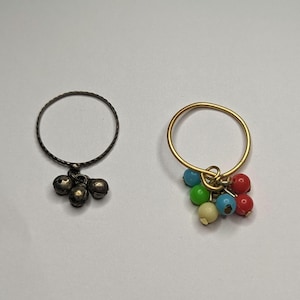 May include: Two rings with charms. The first ring is black and has three small bells hanging from it. The second ring is gold and has a cluster of colorful beads hanging from it.