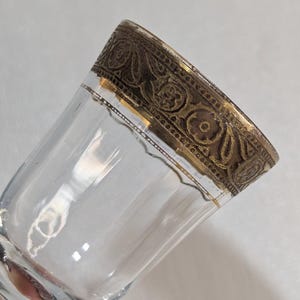 May include: Clear glass tumbler with a decorative, ornate gold-colored rim. The rim features a detailed floral and scroll design. The glass is slightly angled, showcasing its transparency and the intricate detailing of the rim.