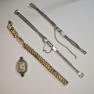 May include: Three watch bands, one gold and two silver, on a white background.