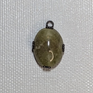 May include: Oval-shaped pendant with a green stone and a dark metal bail. The stone has a mottled appearance with shades of green and brown. The pendant is accented with dark metal details on the sides.