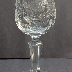 May include: Clear wine glass with etched grape and leaf design. The glass has a rounded bowl, a faceted stem, and a circular base. The etched design covers the bowl of the glass. The glass is made of clear glass.