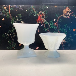 May include: Two white milk glass pedestal bowls, one tall and one short, with a textured design. The bowls are on a white surface.