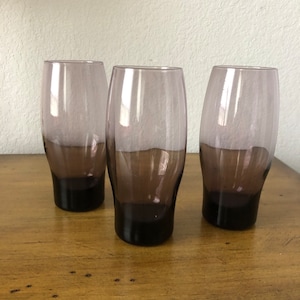 May include: Three purple glass tumblers with a gradient from light to dark purple. The glasses have a rounded bottom and a straight top.