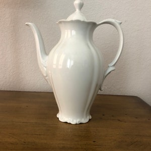 May include: A white porcelain teapot with a lid and a curved spout. The teapot has a fluted body and a decorative handle. The base of the teapot has a scalloped edge. The teapot is sitting on a wooden surface.