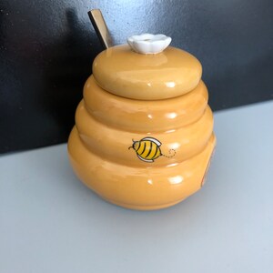 May include: A yellow ceramic honey pot shaped like a beehive with a white flower on the lid and a bee design on the side. The pot has a small spoon attached to the lid.