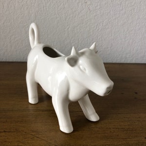 May include: White ceramic cow creamer with a spout. The cow is standing on all four legs and has a hole in the back for pouring.