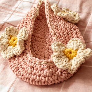 May include: A handmade, pink crochet bag with three large, cream-colored flowers with yellow centers. The bag is in a teardrop shape and sits on a pink plaid surface. The flowers are attached to the front and side of the bag.
